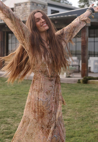 A woman with long brown hair wearing a flowing beige and brown patterned bohemian maxi dress