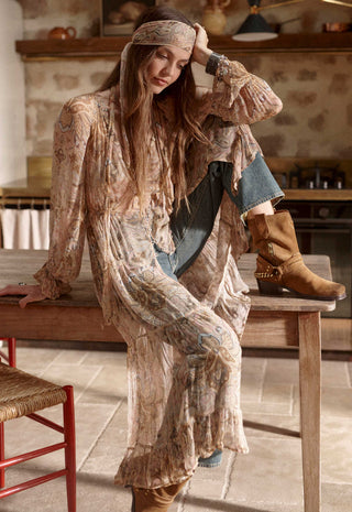 A woman in a bohemian-style room wearing a long, loose beige and brown patterned tunic dress with a matching headscarf, blue jeans, and tan boots, sitting on a wooden table.