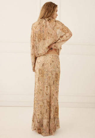 A woman stands with her back to the camera wearing a matching two-piece set consisting of a long-sleeve cropped top and a long skirt, both in a beige and brown paisley patterned fabric.