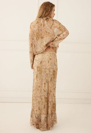 A woman stands with her back to the camera wearing a matching two-piece set consisting of a long-sleeve cropped blouse and a long skirt, both in a beige paisley pattern with brown and muted gold details.