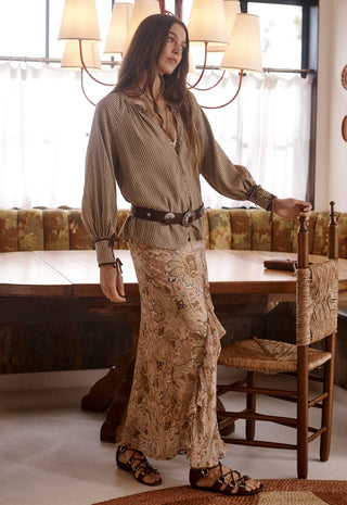 A woman stands in a rustic dining room wearing a long-sleeve beige patterned blouse with a black belt and a matching long skirt with a paisley print. She is wearing brown strappy sandals and leaning on a wooden chair next to a table.