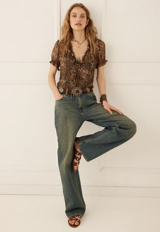 A woman posing in a brown patterned short-sleeve blouse, blue wide-leg jeans, and brown strappy sandals against a white paneled wall.