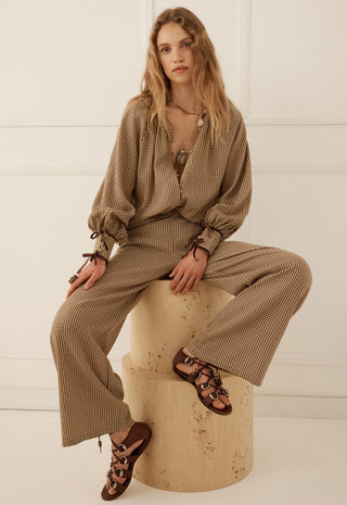 A woman sits on a round stone pedestal wearing a brown and beige gingham two-piece set consisting of a long-sleeve blouse and wide-leg trousers, paired with brown strappy flat sandals against a white paneled wall.