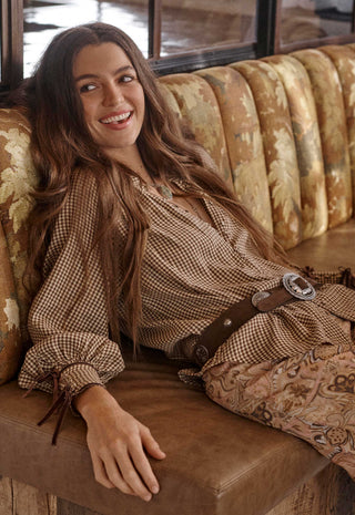 A woman with long brown hair reclines on a brown leather bench wearing a brown and beige gingham long-sleeve top with a matching brown belt and patterned pants