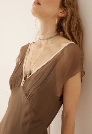 A close-up lifestyle shot of a model wearing a brown semi-sheer draped dress with a deep V-neckline and layered silver necklaces.